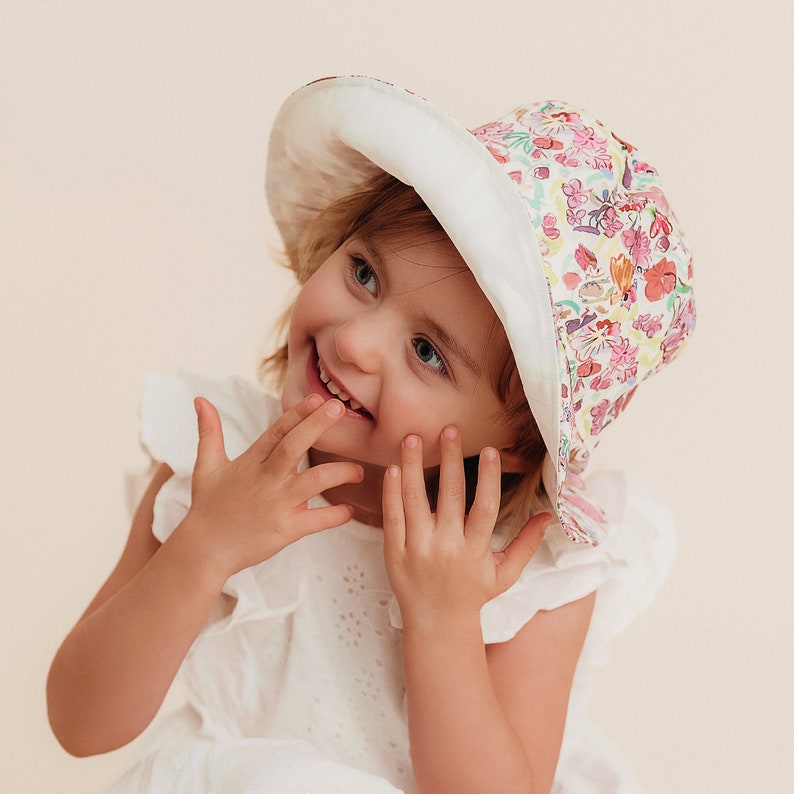 May include: A young girl wearing a white dress with a floral print sun hat. The hat has a white brim and a colorful floral pattern.