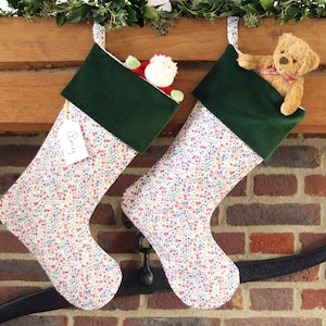 May include: Two Christmas stockings with a floral pattern of red and blue flowers on a cream background, topped with green velvet. One stocking has a tag that says "Barry". One stocking is filled with a Santa Claus toy, and the other has a teddy bear.