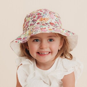 May include: A young girl wearing a white ruffle top and a floral print sun hat with a wide brim. The hat has a pink, yellow, and green floral pattern.