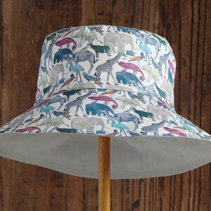 May include: A white bucket hat with a colorful print of various animals, including elephants, giraffes, and birds. The hat has a wide brim and a white underside.