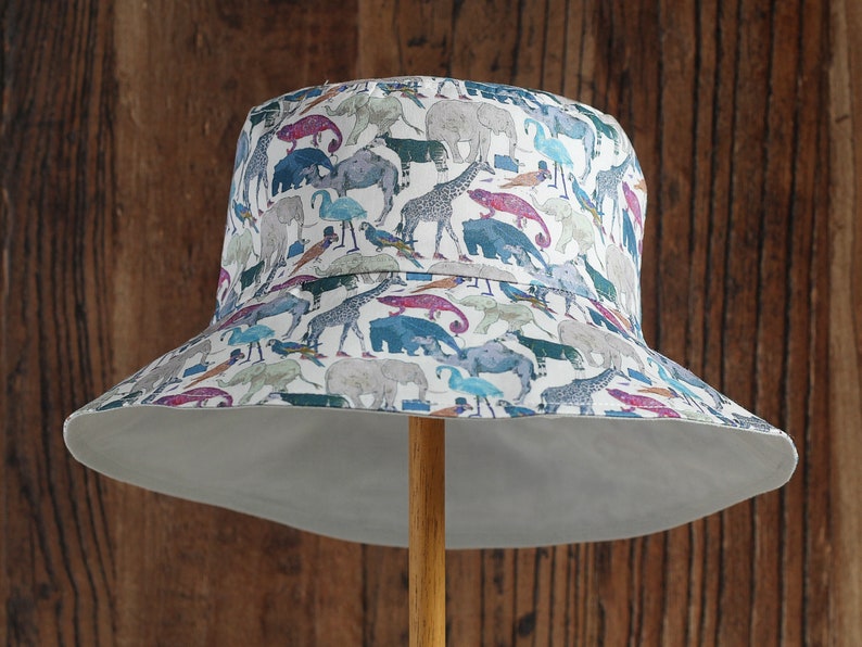 May include: A white bucket hat with a colorful print of various animals, including elephants, giraffes, and birds. The hat has a wide brim and a white underside.