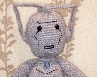 May include: A gray crocheted plush toy in the shape of a robot with a blue button on its chest. The robot has a large head with a wide, flat face and large, black eyes.