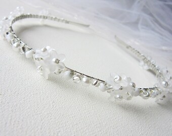 May include: A bridal headband with white flower and pearl detailing. The floral design is set on a thin, silver-coloured wire frame. The image shows a sheer white veil in the background.