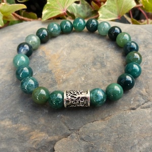 Op de afbeelding: Een armband met ronde, gepolijste groene kralen en een zilverkleurig cilindrisch accent. De armband ligt op een grijze stenen ondergrond, met groen blad in de achtergrond. De kralen variëren in groentinten.