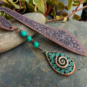 May include: Copper bookmark with an ornate, engraved design. The bookmark features a teardrop-shaped charm with a teal spiral design and teal beads. The bookmark is resting on a grey stone.