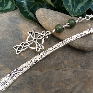 Puede incluir: Un marcapáginas de metal plateado con un patrón floral en relieve a lo largo de su longitud. En la parte superior lleva un dije de cruz celta plateada y dos cuentas esféricas de color verde oscuro separadas por pequeños espaciadores plateados.