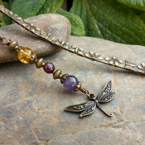 Peut inclure: Un marque-page en laiton avec un motif floral et un charme de libellule. Le marque-page est orné de perles dans les tons ambre, violet et grenat.