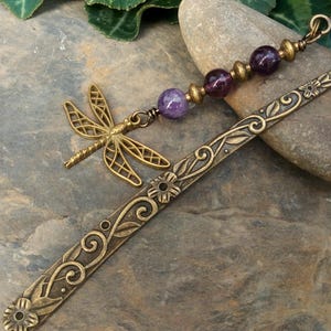 Può includere: Un segnalibro in metallo color bronzo con un motivo floreale e a viticci in rilievo. Sulla parte superiore è fissato un ciondolo a forma di libellula e una serie di tre perle di pietra levigata viola e rosso scuro separate da distanziatori dorati.