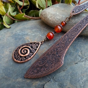 May include: A copper bookmark and pendant set featuring detailed floral and spiral patterns. The bookmark has a curved handle, and the pendant is teardrop-shaped. Both are linked by a chain with orange beads. The set is presented on a grey stone surface.