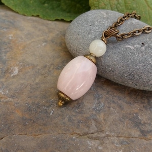Op de afbeelding: Een ketting met een bronzen ketting en roze en witte edelstenen. De hanger heeft een grote ovale roze edelsteen en een kleinere witte edelsteen.
