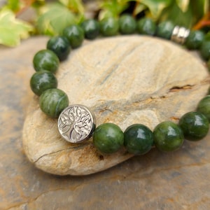 Puede incluir: Una pulsera de cuentas verdes con un cierre plateado con un diseño de árbol. La pulsera está hecha de cuentas verdes redondas y pulidas. La pulsera descansa sobre una piedra de color marrón claro, con follaje verde en el fondo.