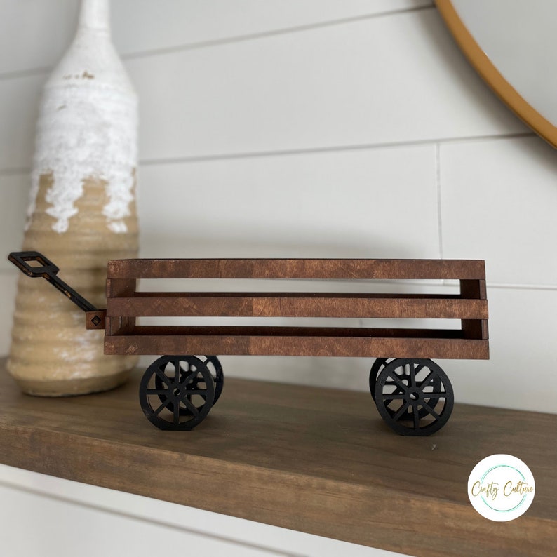 May include: A small wooden wagon with black metal wheels and a handle. The wagon has two shelves and is stained a dark brown color. The wagon is sitting on a wooden shelf.