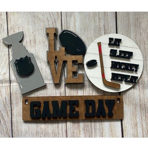 May include: Wooden sign with the words "LOVE" and "GAME DAY" in black letters. The sign also includes a hockey stick, puck, and a trophy with the words "EAT SLEEP HOCKEY REPEAT" on a white circle.