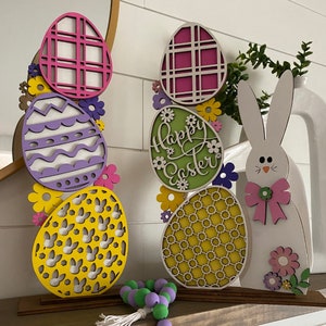 May include: A set of two wooden Easter decorations, each featuring a stack of colorful, laser-cut eggs with a different pattern. The left decoration has a purple egg with a wavy pattern, a yellow egg with a bunny pattern, and a pink egg with a checkered pattern. The right decoration has a green egg with the words "Happy Easter" in a script font, a yellow egg with a honeycomb pattern, and a pink egg with a checkered pattern. Both decorations are accented with small, colorful flowers.