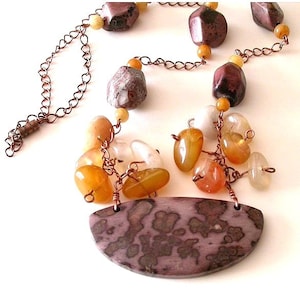 May include: A long copper chain necklace with a large, brown and white agate pendant. The pendant is shaped like a half-moon and has a mottled pattern. The necklace is adorned with smaller agate beads in shades of brown, orange, and white.