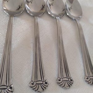 May include: Four stainless steel spoons with a reflective surface. The spoon handles have a ribbed design that widens towards the bottom, with a decorative flourish at the base. The spoons are arranged on a white surface.