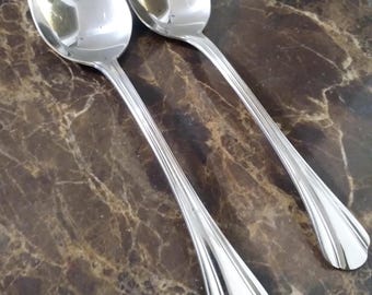 May include: Two shiny, silver spoons with oval bowls and long, slender handles. The handles have a decorative, ridged design near the bowl. The spoons are resting on a brown and tan marble-patterned surface.