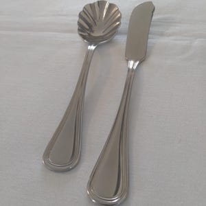 May include: A stainless steel butter knife and a small spoon with a shell-shaped bowl. The spoon and knife have a simple, elegant design with a polished finish. The utensils are likely for serving or eating.