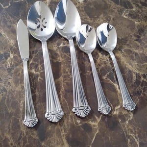 May include: A set of five silver-colored serving utensils, including a butter knife, a slotted spoon, a serving spoon, and two smaller spoons. The utensils have a decorative, ridged design on the handles and are arranged on a brown marble surface.