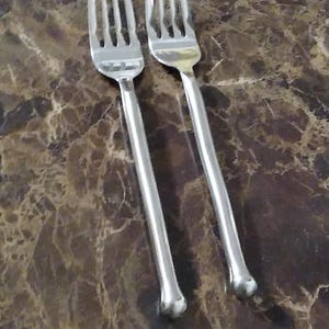 May include: Two stainless steel forks with long, slender handles and four tines each. The forks are positioned diagonally against a brown and tan marble-patterned surface, creating a simple, elegant composition. The handles have a rounded, decorative end.