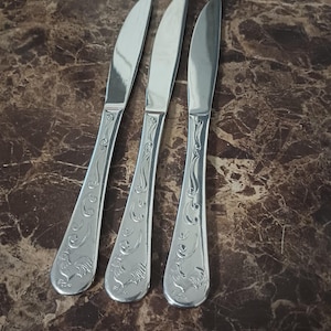 May include: Three silver butter knives with decorative handles featuring a rooster design. The knives have a curved blade and are arranged on a brown marble-patterned surface. The handles have an ornate, engraved pattern.