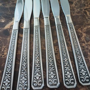 May include: A set of six silver-toned stainless steel dinner knives arranged in a row. Each knife features a smooth, polished blade and a flat handle decorated with an embossed, repeating floral and scroll pattern in a dark, oxidized finish.