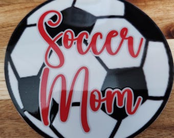 May include: A white and black soccer ball with the words "Soccer Mom" in red cursive lettering.