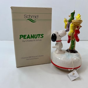 May include: A ceramic Snoopy figurine from the Peanuts series, with a red collar, stands on a snowy base. A Christmas tree with hanging stockings is behind Snoopy. The box reads "PEANUTS by Charles M. Schulz" and "Christmas 1980 Musical Collectible".