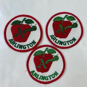 Könnte beinhalten: Drei runde Aufnäher mit einem roten Apfeldesign, grünen Blättern und einer grünen Raupe. Der Apfel hat einen grünen Buchstaben "A" und das Wort "ARLINGTON" in grüner Schrift. Die Aufnäher haben einen weißen Hintergrund und einen roten Rand.