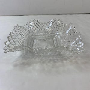 May include: Clear glass square bowl with a diamond pattern and a ruffled edge. The bowl has a square base with a starburst design in the centre. The edges of the bowl are scalloped.