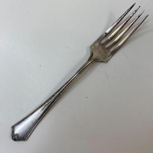 May include: A silver fork with four tines, featuring a decorative handle. The fork has a slightly tarnished appearance, suggesting age or use. The handle has a detailed design near the base.