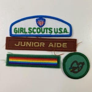 Pode incluir: Uma coleção de patches Girl Scouts USA. Os patches incluem um patch arqueado azul e branco com o texto "GIRL SCOUTS USA", um patch retangular marrom com "JUNIOR AIDE", um patch listrado arco-íris e um patch circular verde.