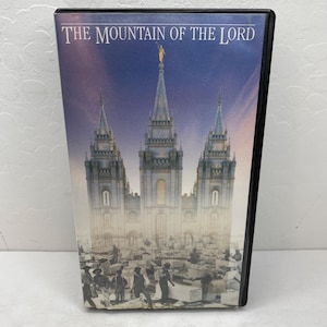 May include: A DVD case featuring a detailed illustration of a tall, ornate building with multiple spires, set against a blue and purple sky. The text "THE MOUNTAIN OF THE LORD" is displayed at the top. The bottom of the image shows figures and stone blocks.
