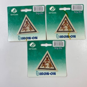 Może przedstawiać: Trzy naszywki termoprzylepne Try-Its dla dziewcząt z harcerstwa. Każda naszywka jest trójkątna z beżowym tłem, brązową obwódką i wzorem trzech niedźwiedzi trzymających serca. Naszywki są na zielonych kartach.