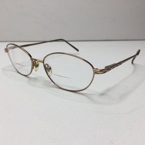 Könnte beinhalten: Eine ovale Brille mit einem braunen Metallrahmen. Der Rahmen hat ein geflochtenes Design an den Bügeln.