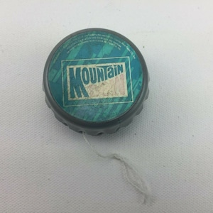 Puede incluir: Un yoyo gris con una cuerda blanca. El yoyo tiene una etiqueta azul y verde con el texto "Mountain Dew" en ella.