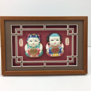 Puede incluir: Una obra de arte enmarcada con dos figuritas coloridas de niños que sostienen peces. Las figuritas están colocadas sobre un fondo rojo con un borde decorativo. La obra de arte está enmarcada en un marco de madera marrón.