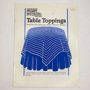 May include: A vintage book cover titled "Table Toppings" by Instant Interiors. The cover features a blue and white graphic of a tablecloth. The text reads, "Create Your Own Tablecloths, Napkins, Placemats, Runners."