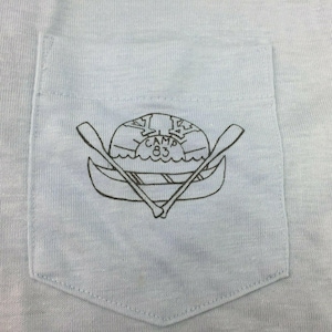 May include: A light blue t-shirt with a pocket featuring a black and white illustration of a canoe with two paddles and the text "Camp 83".