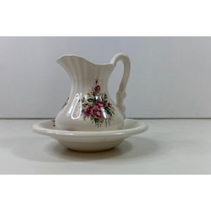 May include: A white ceramic pitcher and basin set. The pitcher has a curved handle and a flared spout. Both pieces are decorated with a floral pattern of pink roses and green leaves. The set is designed for decorative use.