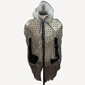 May include: A clear, hooded raincoat with black polka dots and black trim. The jacket has a zipper closure, two front pockets, and a drawstring hood. The raincoat is displayed on a mannequin.