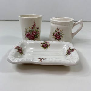 May include: A three-piece bathroom set featuring a white ceramic tumbler, mug, and soap dish. Each piece is adorned with a floral rose pattern in shades of pink and green. The soap dish is rectangular with a decorative edge.