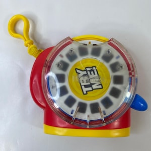 Pode incluir: Um brinquedo View-Master vintage em plástico vermelho, amarelo e transparente. O visor circular tem um centro amarelo com o texto "View-Master". Uma alavanca azul e um laço amarelo estão anexados. O visor exibe pequenas imagens retangulares.