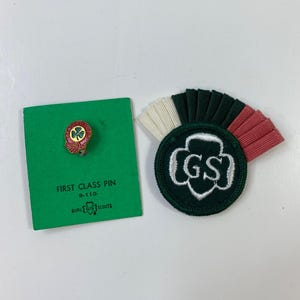 May include: A green card with a First Class Pin and Girl Scout logo, next to a dark green patch with white embroidered "GS" letters and a fan of white, dark green, and red fabric.
