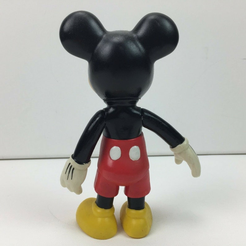 May include: A black and red plastic Mickey Mouse figurine. The figurine is standing with its arms outstretched and is wearing red shorts with white circles. The figurine has black ears and yellow shoes.