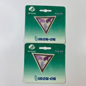 Puede incluir: Dos parches termoadhesivos Girl Scouts Try-Its. Cada parche presenta un triángulo con tres manos moradas y una mano morada clara en el centro. Los parches están sobre tarjetas de respaldo verdes con las palabras "Girl Scouts", "Try-Its" e "IRON-ON".