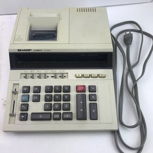 May include: A vintage Sharp Compet CS-2606 calculator with a built-in printer. The calculator has a beige body with black keys and a red equals key. The text "SHARP COMPET CS-2606" is printed on the front. A gray power cord is attached.