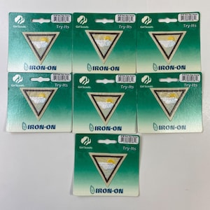 Puede incluir: Varios parches termoadhesivos Try-Its de las Girl Scouts. Cada parche es triangular con un fondo beige, un borde marrón y un diseño de sol amarillo. Los parches están en un embalaje de cartón verde.