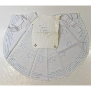 May include: White fabric with blue star embroidery and blue stitched lines. A pattern is on top of the fabric. The fabric has a circular shape with a white strap attached. The pattern has a star design.