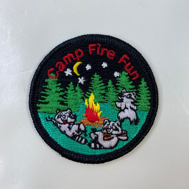 May include: Embroidered patch with a black border and the words "Camp Fire Fun" in red. The patch features three raccoons around a campfire, with a crescent moon, stars, and pine trees in the background. The patch is round.